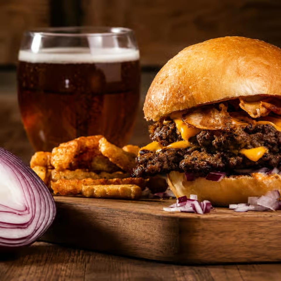 Cheeseburger with waffle fries, red onion, and a glass of beer on a wooden board.