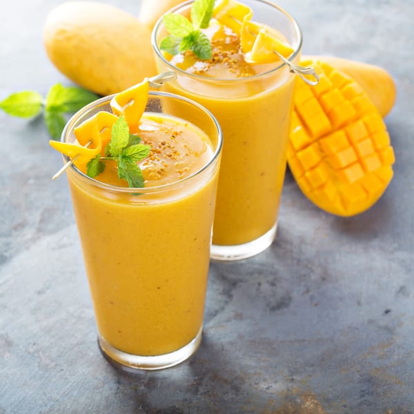 Two mango smoothies garnished with fresh mint and mango slices, with whole and cut mangoes.