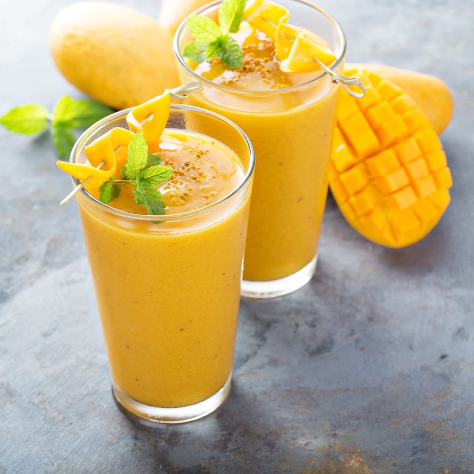 Two mango smoothies garnished with fresh mint and mango slices, with whole and cut mangoes.
