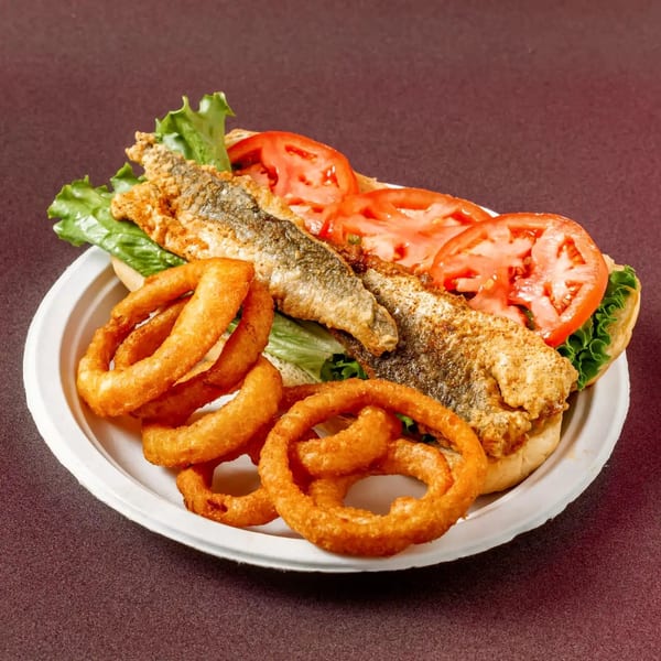 Fried fish sandwich with lettuce, tomatoes, and a side of golden onion rings on a white plate.