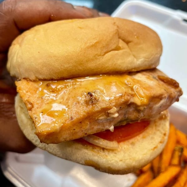 A hand holds a Go To Burgers salmon sandwich with tomato and onion, with fries in the background.